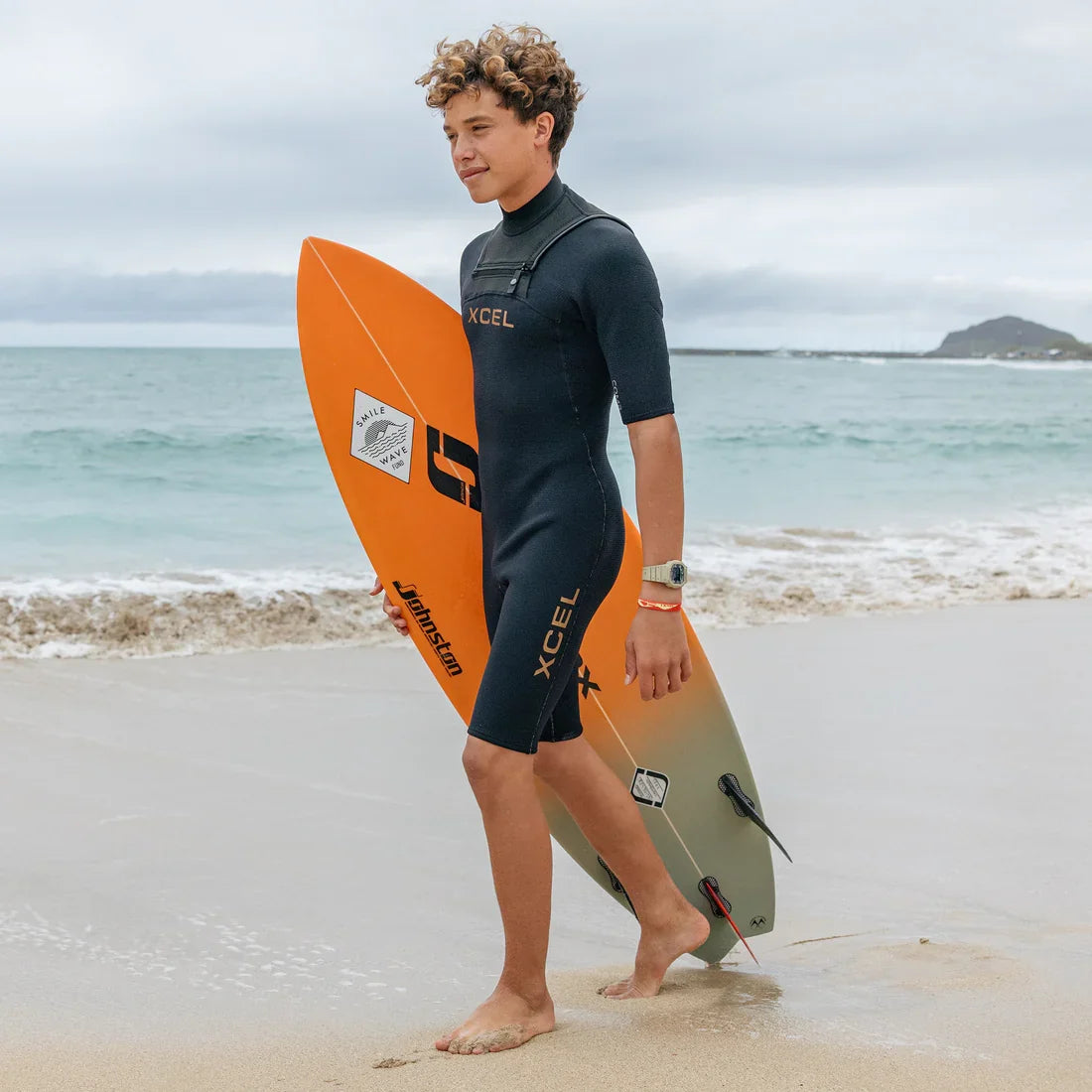 Xcel Mens Comp+ Short Sleeve Springsuit 2/1.5mm worn walking along the beach with a surfboard
